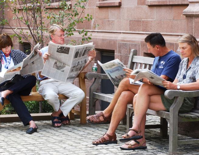 Four people seated on outdoor benches reading newspapers