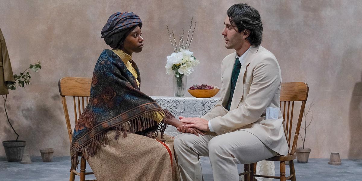Two actors seated at a table on stage