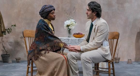 Two actors seated at a table on stage